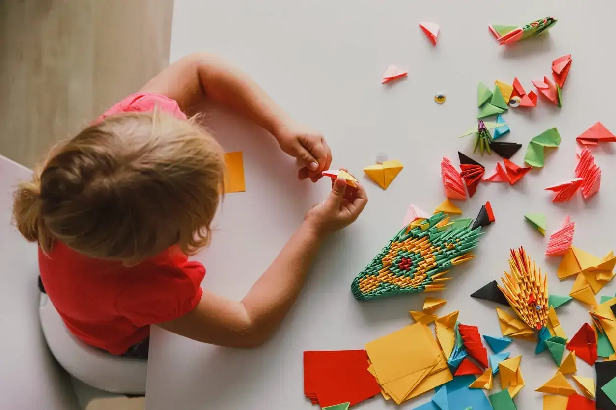 Ni&ntilde;a rubia con camiseta roja crea figuras de papiroflexia espa&ntilde;ol. Dragones y animales de papel decoran la mesa.