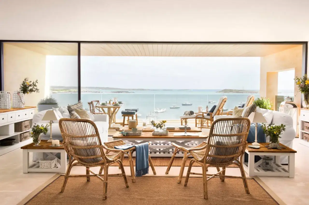 Sal&oacute;n con muebles de mimbre y sof&aacute;s blancos, con una **ventana panor&aacute;mica** que ofrece vistas al mar y barcos.