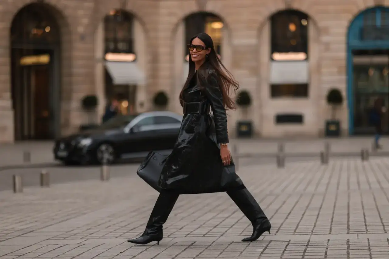 Une femme élégante en trench-coat noir verni, bottes et lunettes de soleil, parfaite pour savoir comment s'habiller quand il fait 10 degrés femme.