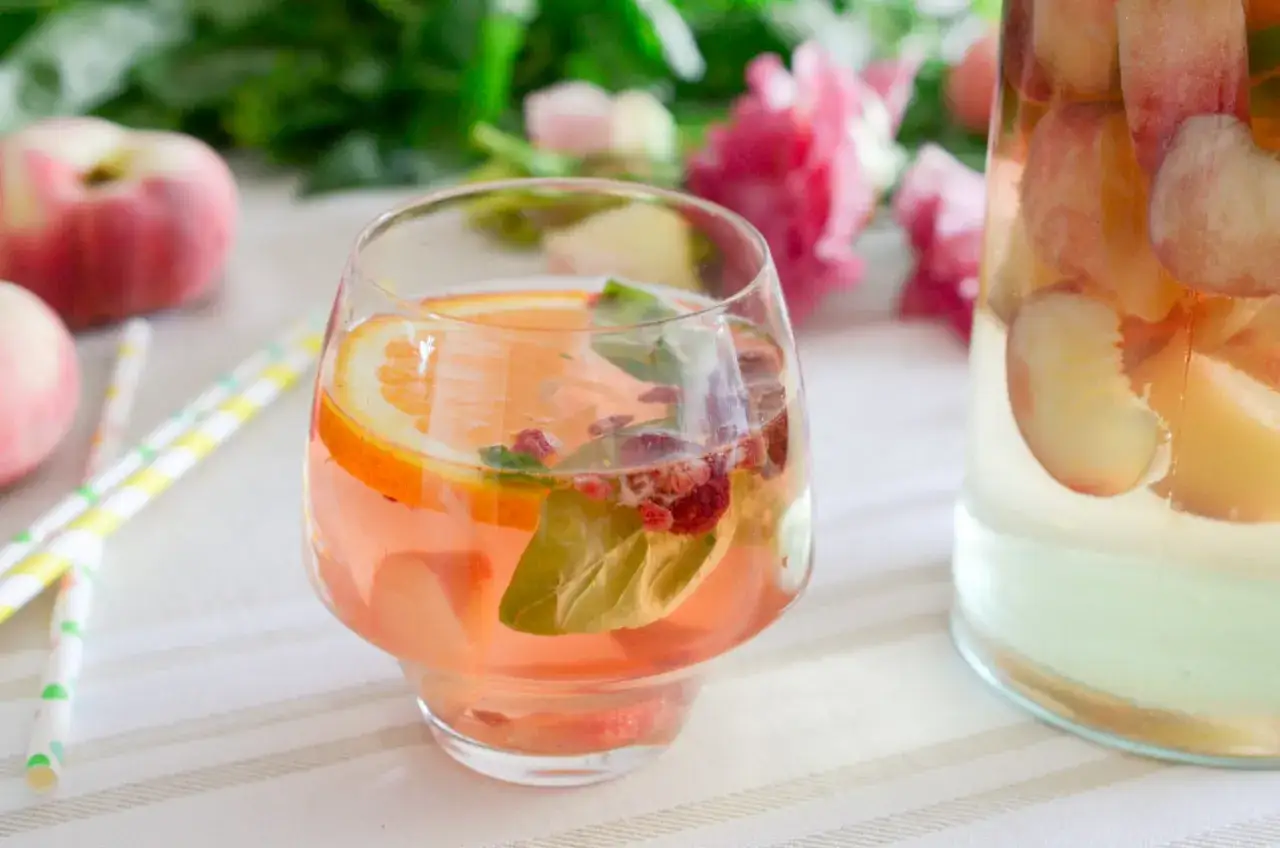 Un verre de sangria blanche aux fruits, avec des tranches d'orange, des framboises et des feuilles de menthe. Une carafe remplie de fruits est visible à droite.