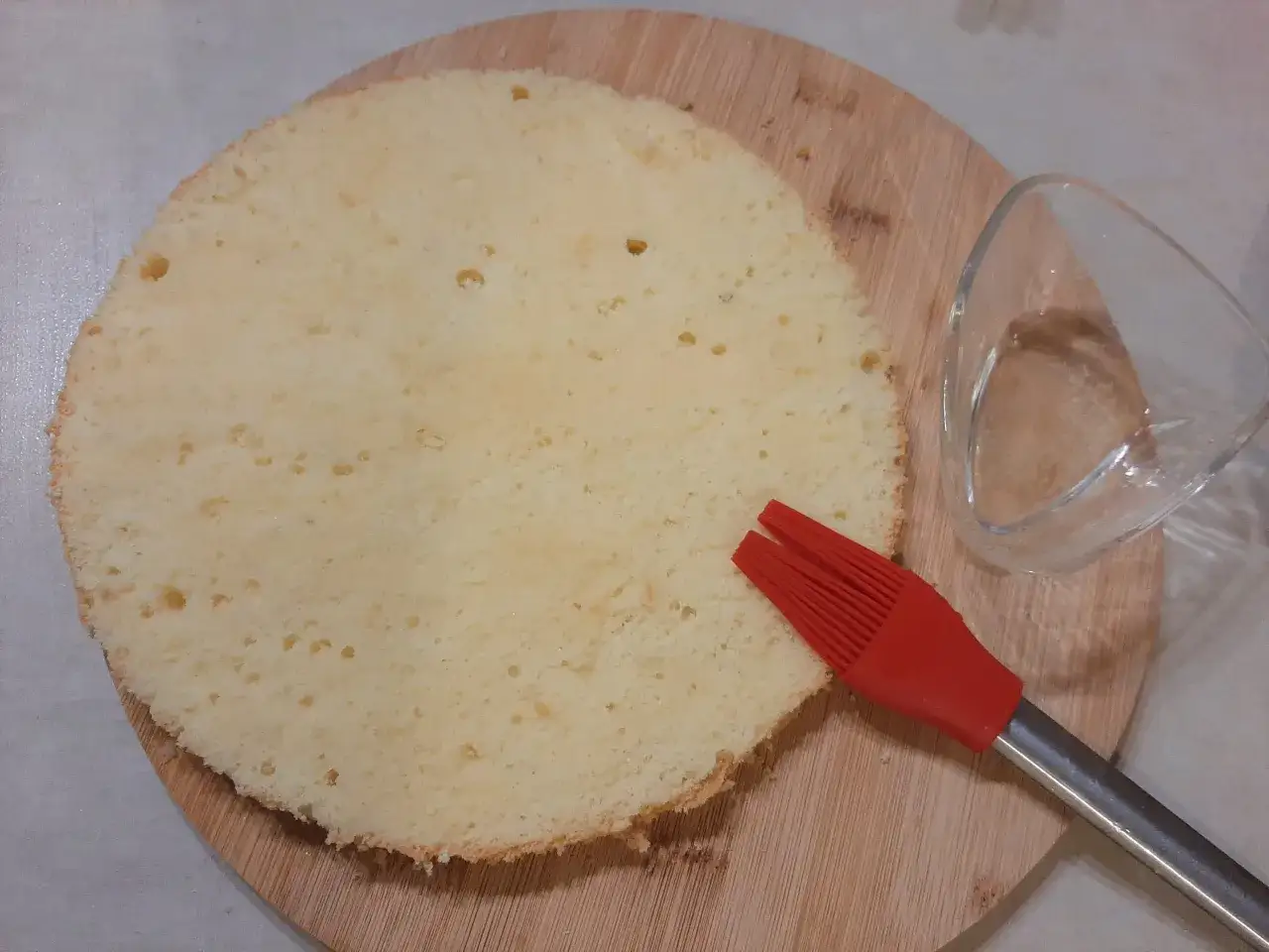 Puszysty biszkopt na desce, obok czerwony pędzelek i szklana miseczka. Czas pomyśleć, czym nasączyć biszkopt, by był wilgotny i pyszny!