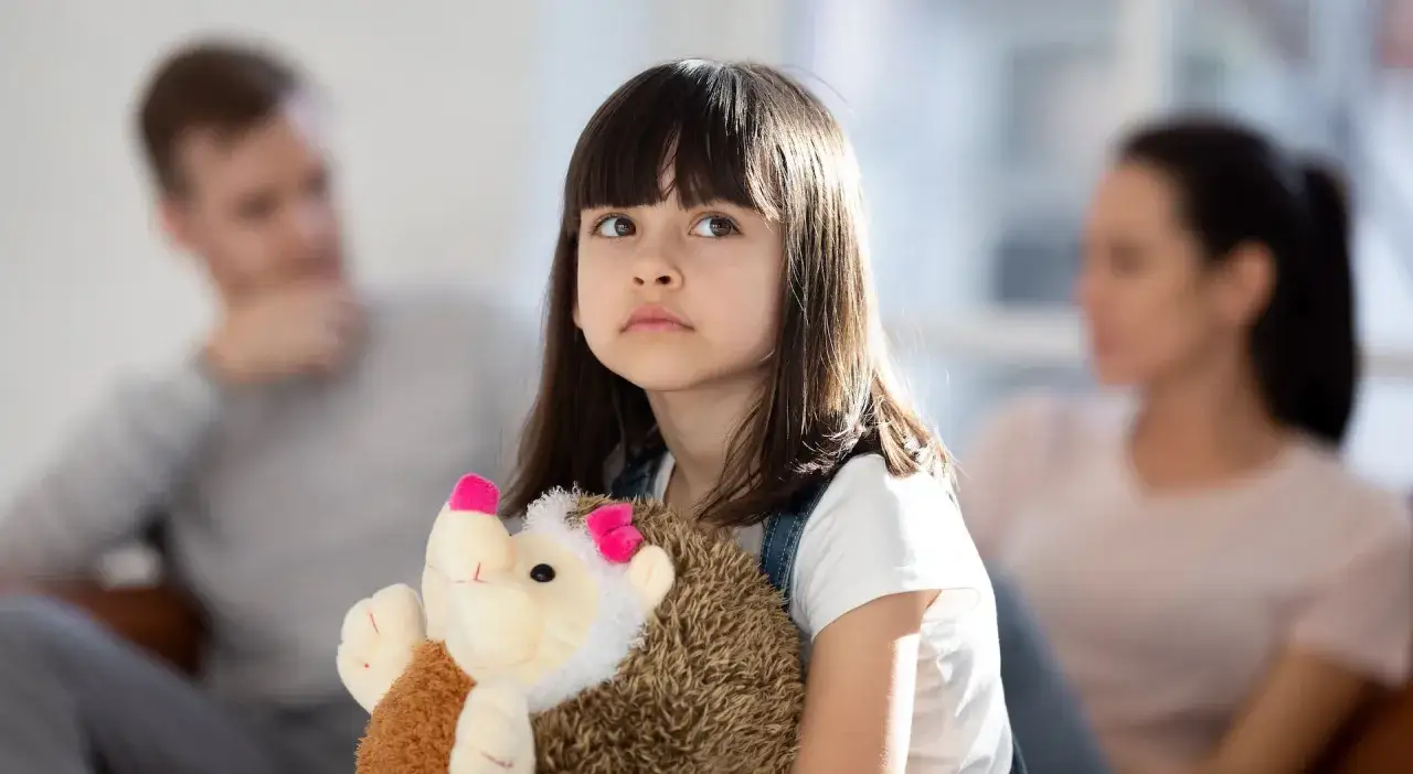 Ein trauriges M&auml;dchen h&auml;lt seinen Teddyb&auml;r fest, w&auml;hrend im Hintergrund die Eltern, die sich trennen, zu sehen sind.