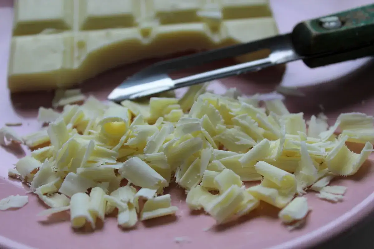 Un économe, accessoire pour faire des copeaux de chocolat, est posé près d'une tablette de chocolat blanc et de ses éclats sur une assiette rose.