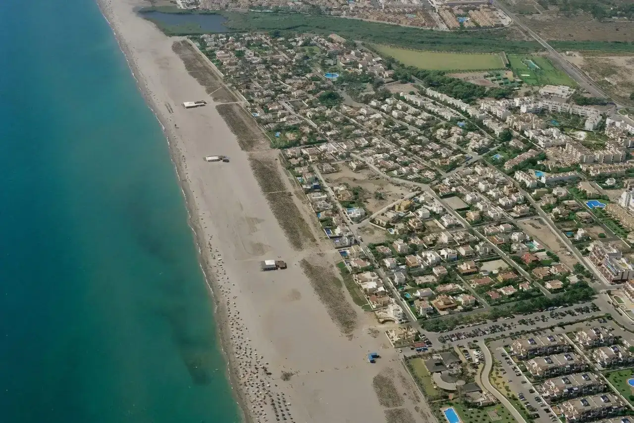 Vista aérea de la playa de Puerto Rey, con el mar turquesa a la izquierda y una extensa zona de arena con sombrillas y algunas construcciones. Detrás, un complejo residencial de casas blancas con tejados marrones.