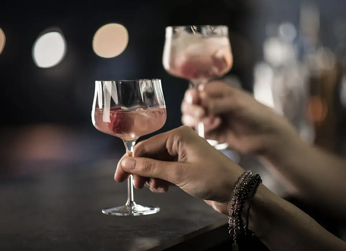 Eleganckie party: poznaj zasady serwowania koktajli rzemieślniczych. Dwie dłonie trzymają kieliszki z różowymi drinkami i malinami.
