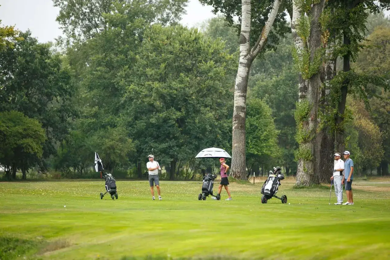 Gracze golfa na polu, wśród drzew. W tle widać torby golfowe i parasol. To może być pierwszy warszawski turniej golfowy.