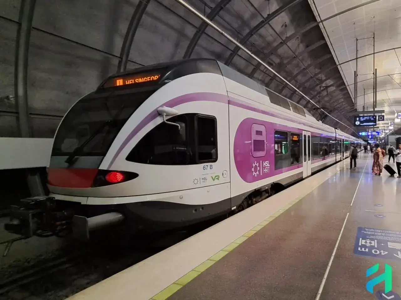 Ein moderner Zug mit lila Akzenten steht am Bahnsteig. Die Anzeige zeigt 