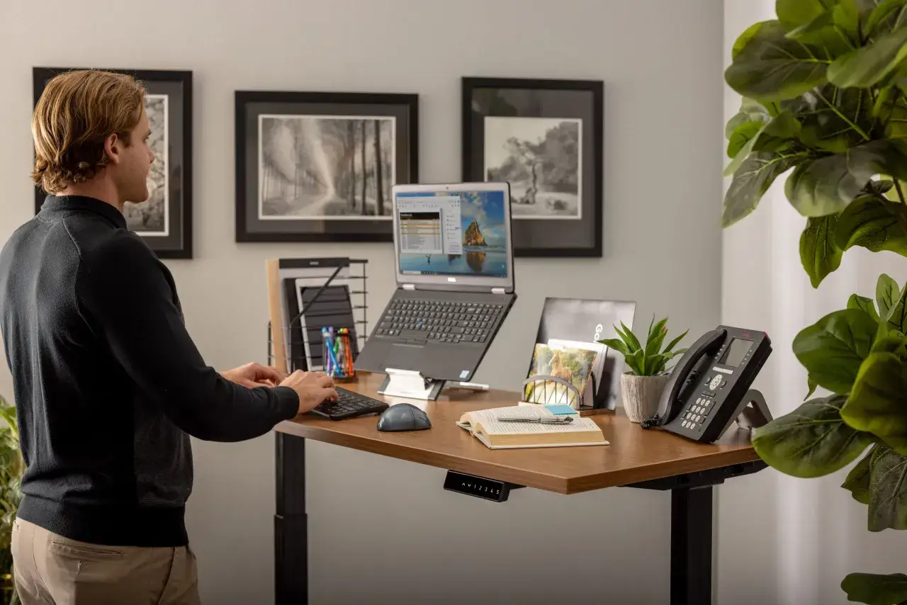 Mann arbeitet an einem Stehschreibtisch mit Laptop. Der laptop arbeitsplatz ist gut organisiert mit Telefon, Pflanzen und Büchern.