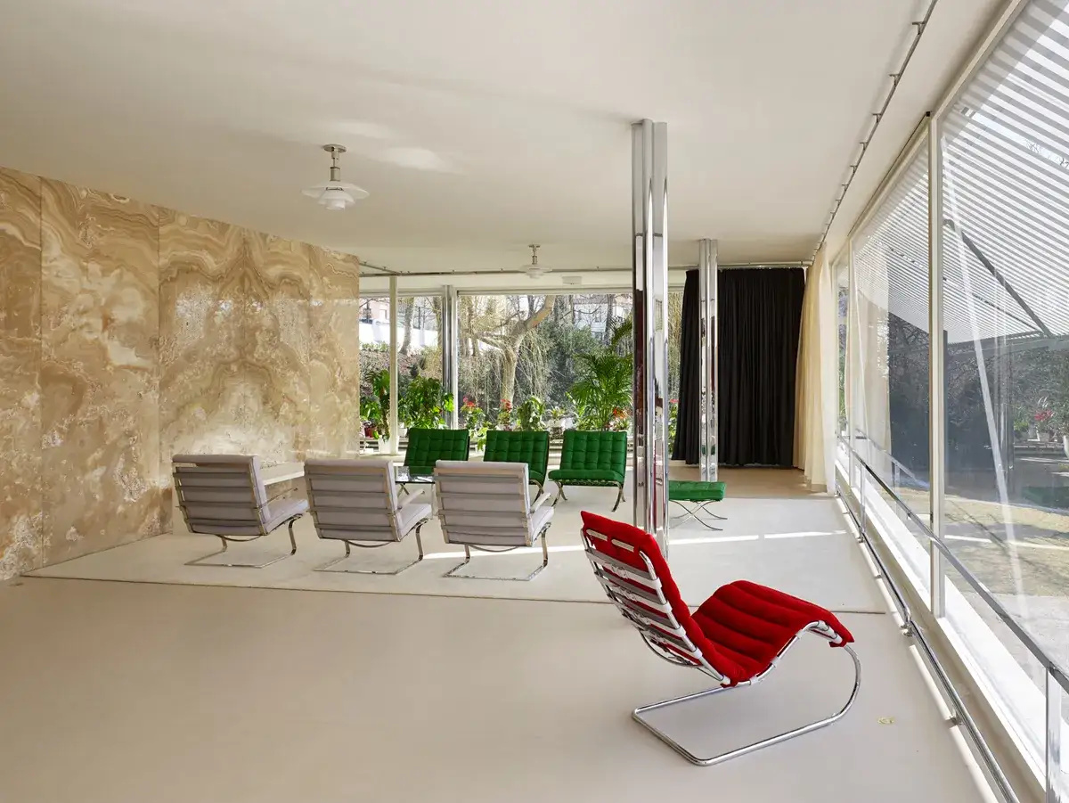 Interno della Casa Tugendhat con poltrone moderne, pareti in travertino e ampie vetrate che si affacciano sul giardino.