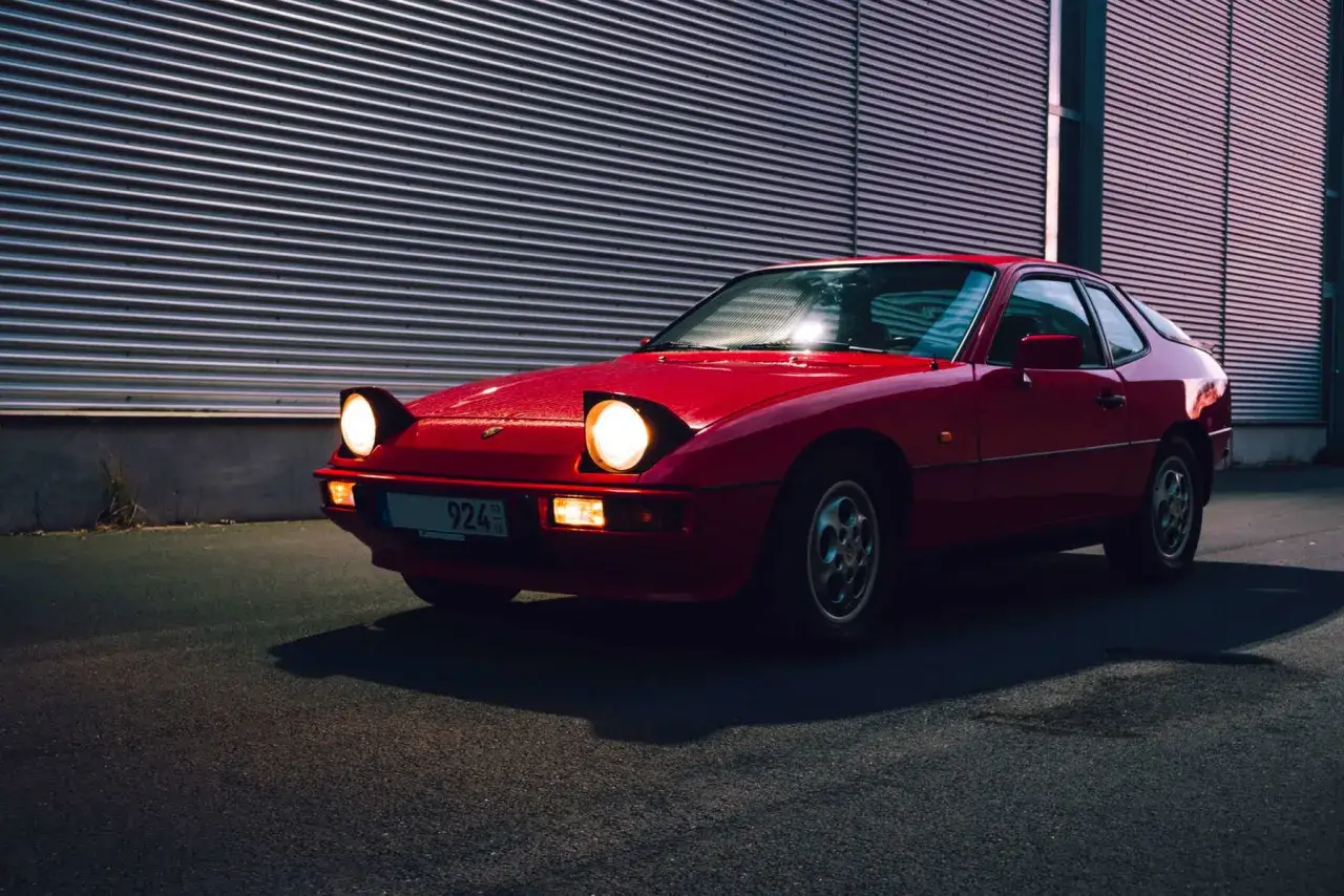Roter Porsche 924 mit ausklappbaren Scheinwerfern vor einer metallischen Wand. Ein Klassiker, dessen **Porsche 924 Verbrauch** auch heute noch &uuml;berrascht.