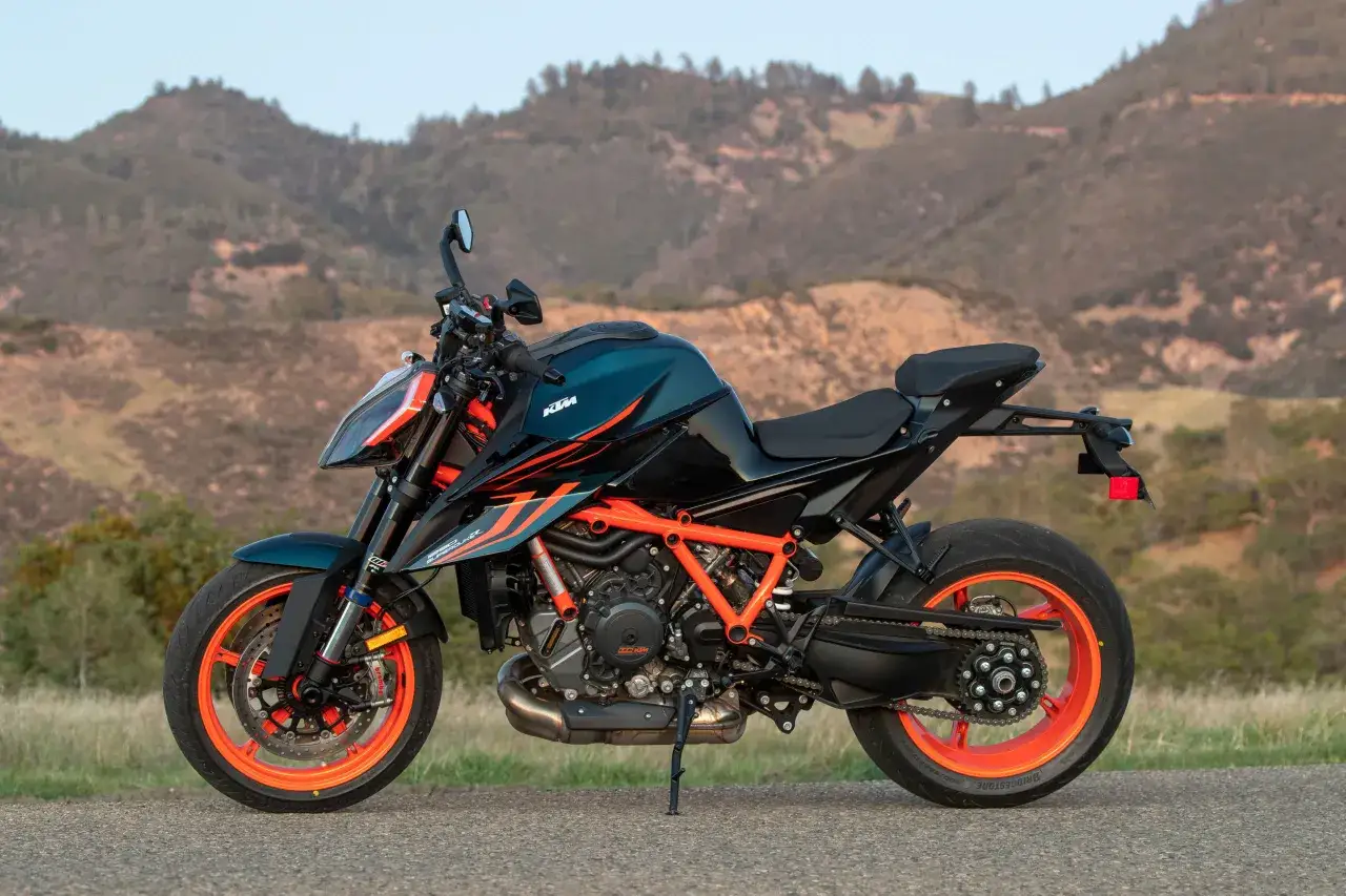 Czarny motocykl KTM Super Duke z pomarańczowymi felgami i ramą, zaparkowany na tle wzgórz.