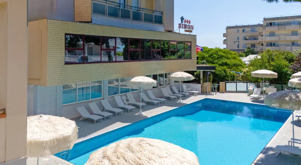 Piscina azzurra e lettini prendisole al **hotel Simon Gatteo Mare**. Ombrelloni di paglia offrono riparo dal sole.