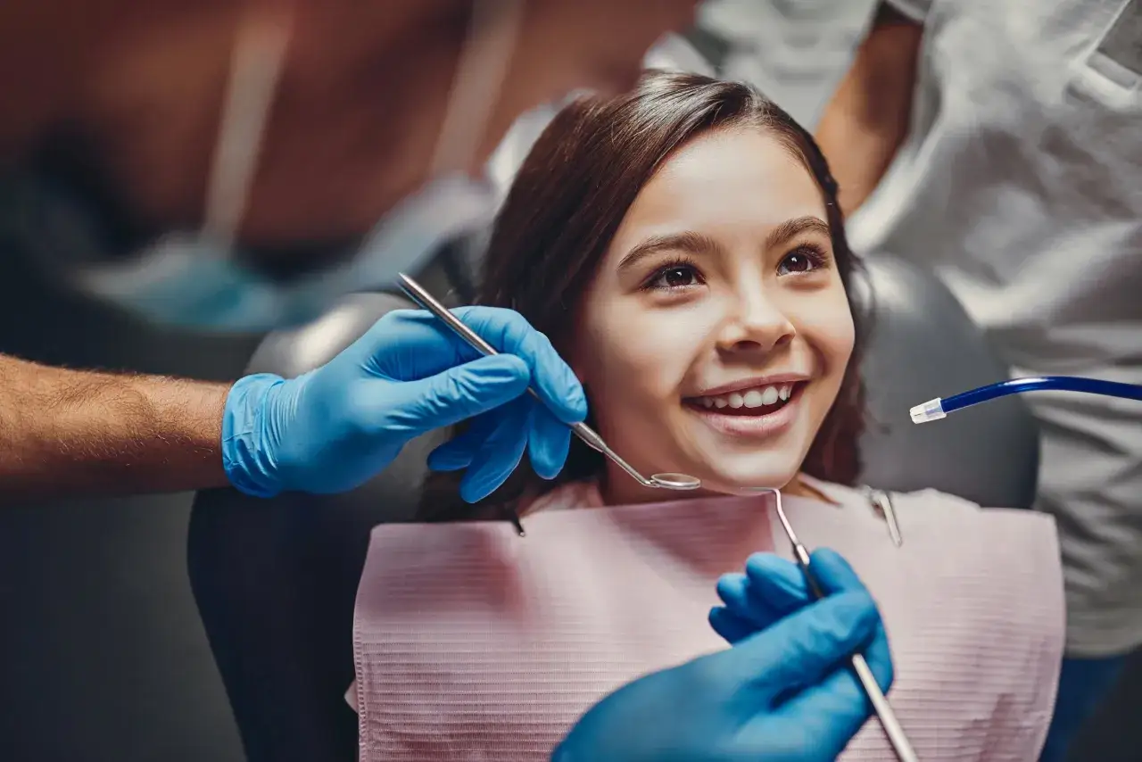 Uśmiechnięta dziewczynka podczas zabiegu lapisowania zębów. Dentysta w niebieskich rękawiczkach używa narzędzi.