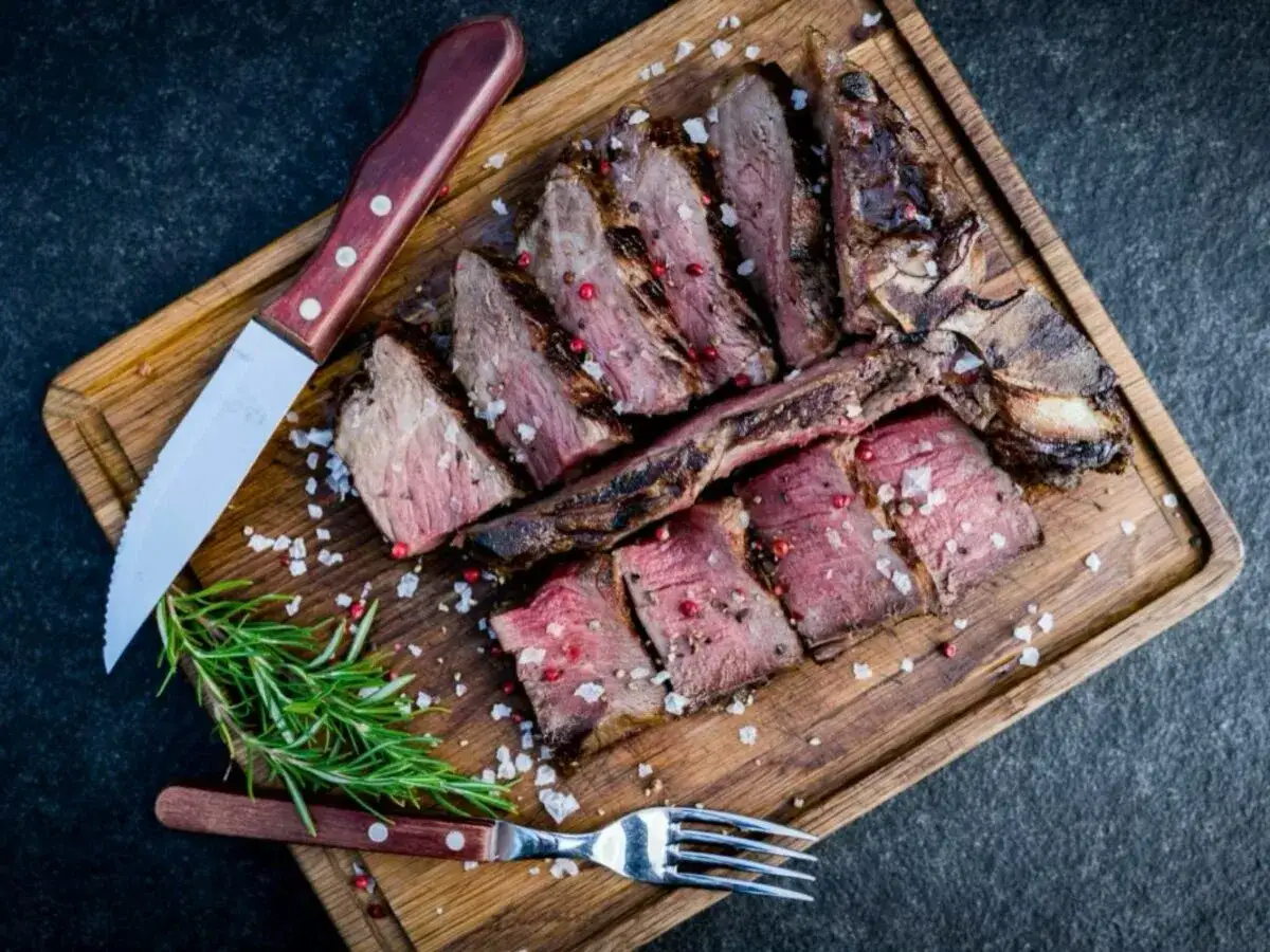 Perfekt gegrilltes Porterhouse Steak, aufgeschnitten und mit grobem Salz und rosa Pfeffer bestreut, liegt auf einem Holzbrett. Ein Messer und eine Gabel liegen daneben.