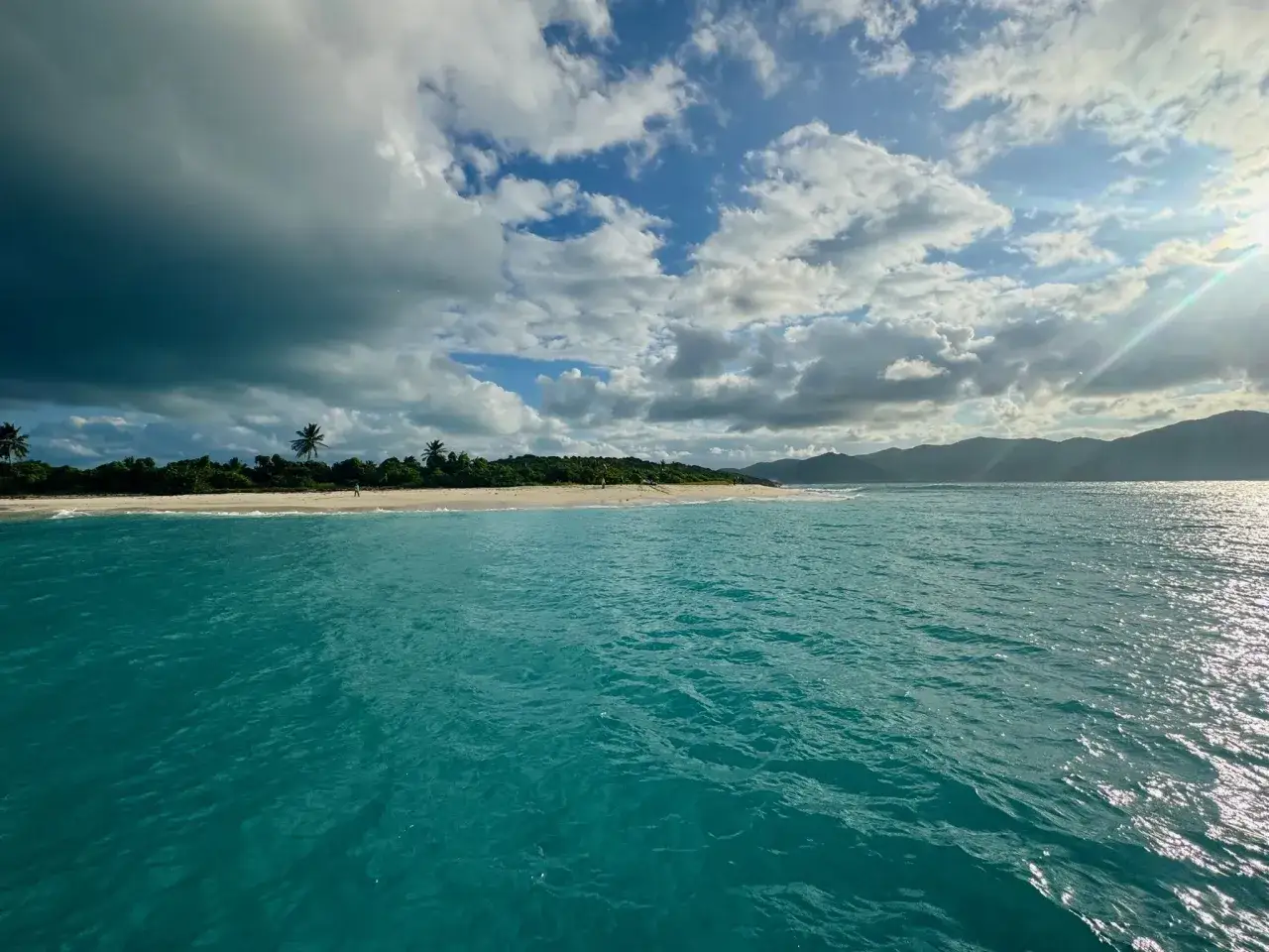 T&uuml;rkisblaues Meer vor einer Insel mit Palmen. Das Wetter auf den Malediven zeigt sich wechselhaft mit dunklen Wolken und Sonnenschein.