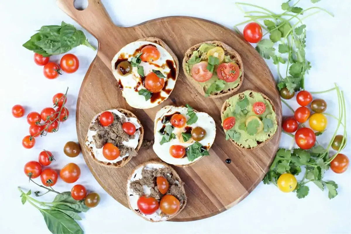 Sju halverade english muffins med olika p&aring;l&auml;gg: &auml;gg, k&ouml;ttf&auml;rs, avokado och tomater. Serveras p&aring; en tr&auml;sk&auml;rbr&auml;da med f&auml;rska &ouml;rter och k&ouml;rsb&auml;rstomater.