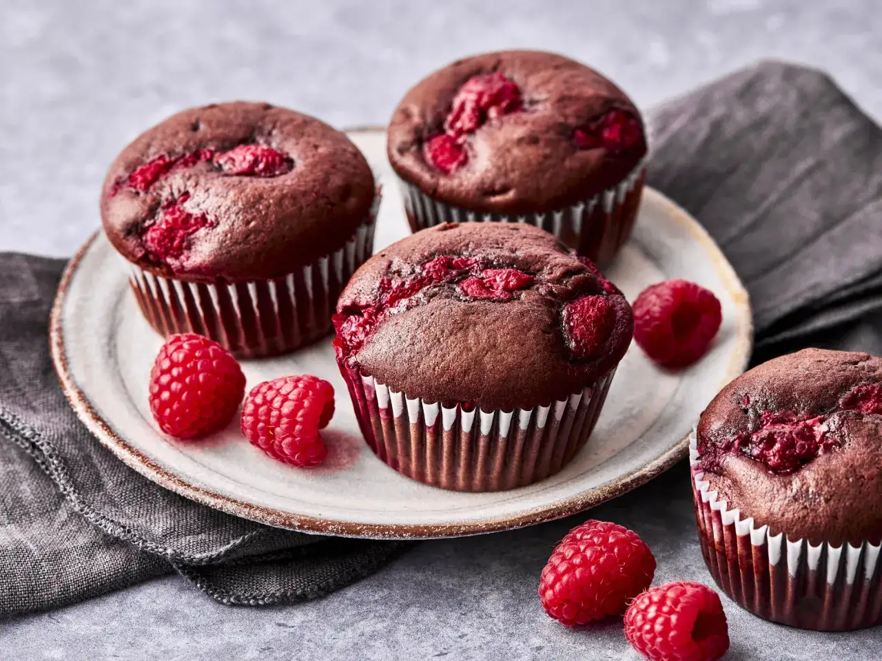 Schokoladige Himbeer-Schoko-Muffins mit frischen Himbeeren auf einem Teller, daneben ein paar lose Himbeeren.