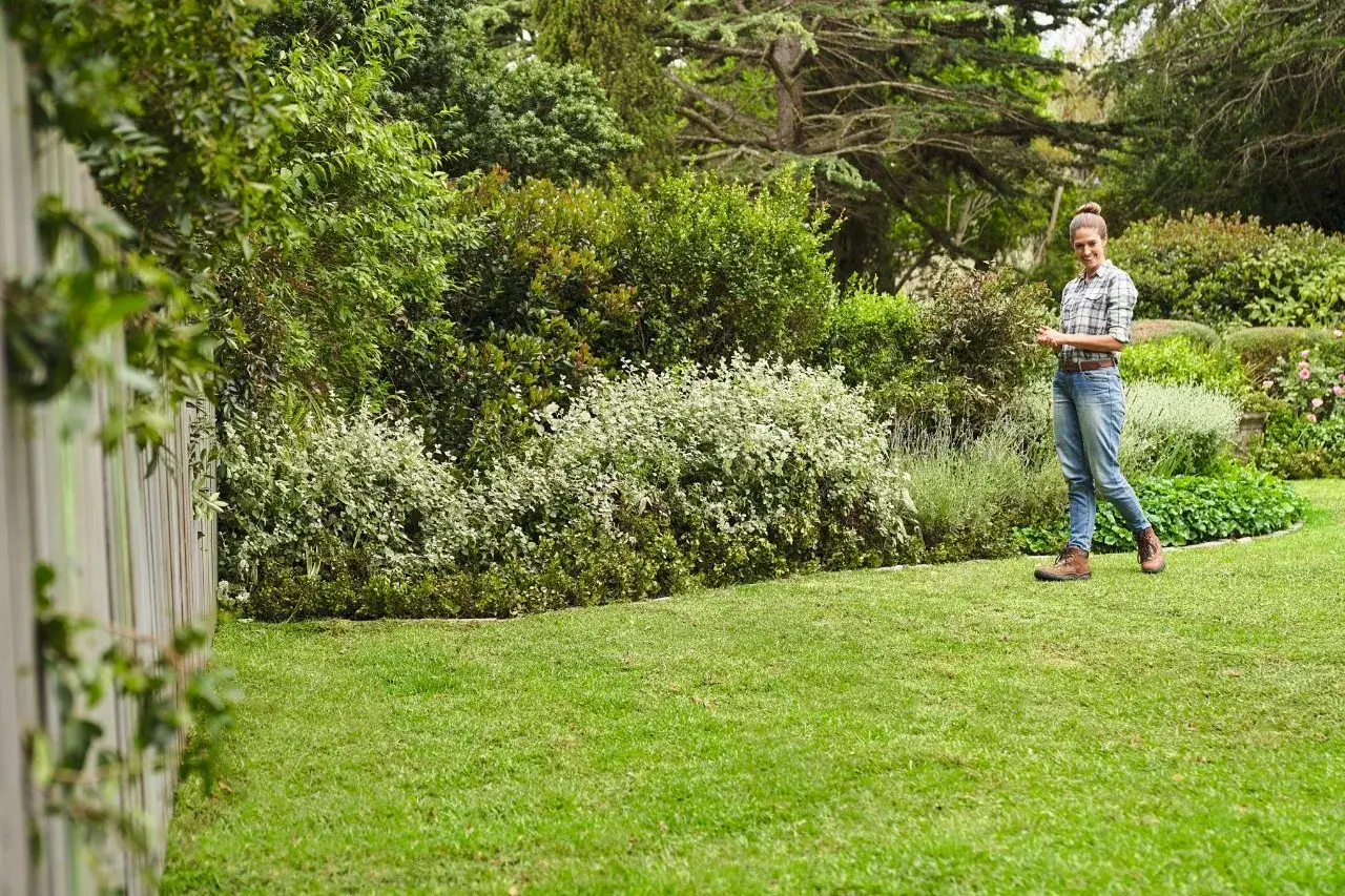 Mujer sonr&iacute;e en un jard&iacute;n exuberante, lista para el cuidado del c&eacute;sped en primavera. C&eacute;sped verde y arbustos floridos.