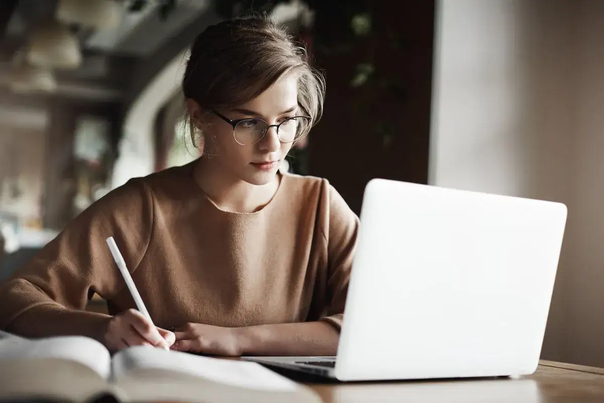 Młoda kobieta w okularach pracuje przy laptopie, pisząc notatki. Idealna scena dla copywriter praca.