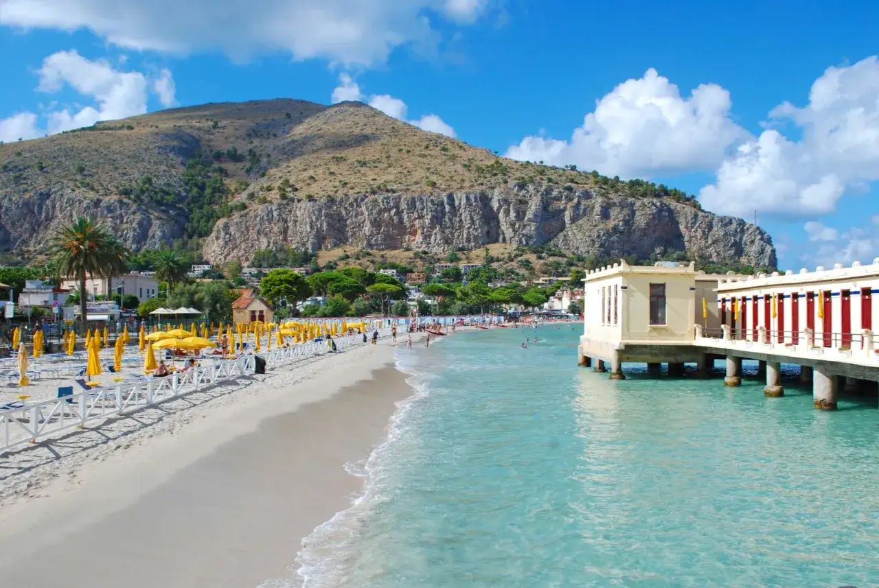 Słoneczna plaża Mondello z turkusową wodą, żółtymi parasolami i górami w tle.