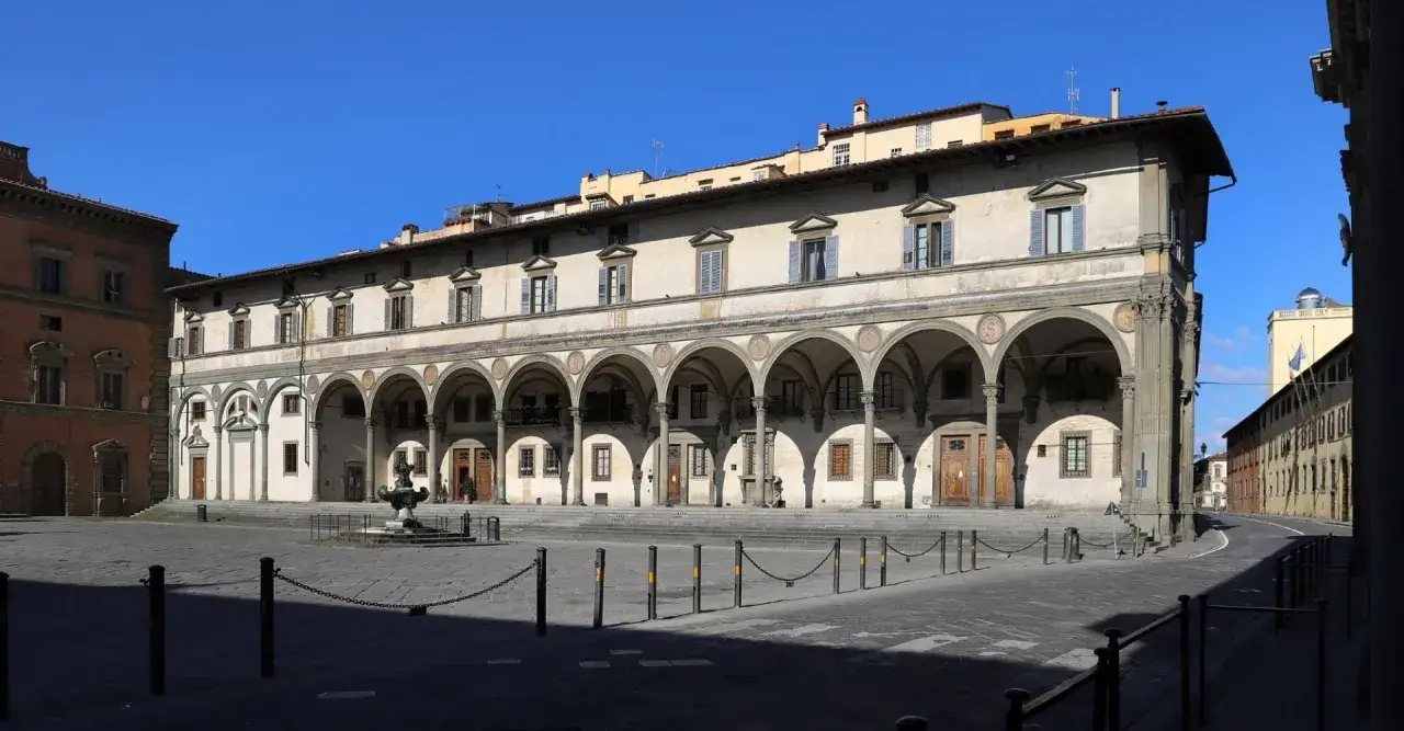 La **piazza della Santissima Annunziata** a Firenze, con il suo portico rinascimentale e la fontana centrale, sotto un cielo azzurro.