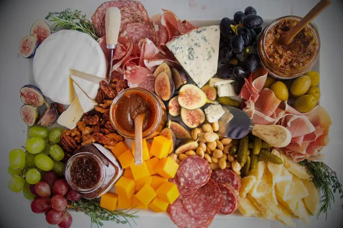 Plateau gourmand avec vin rouge, charcuterie, fromages variés, fruits, noix et confitures.