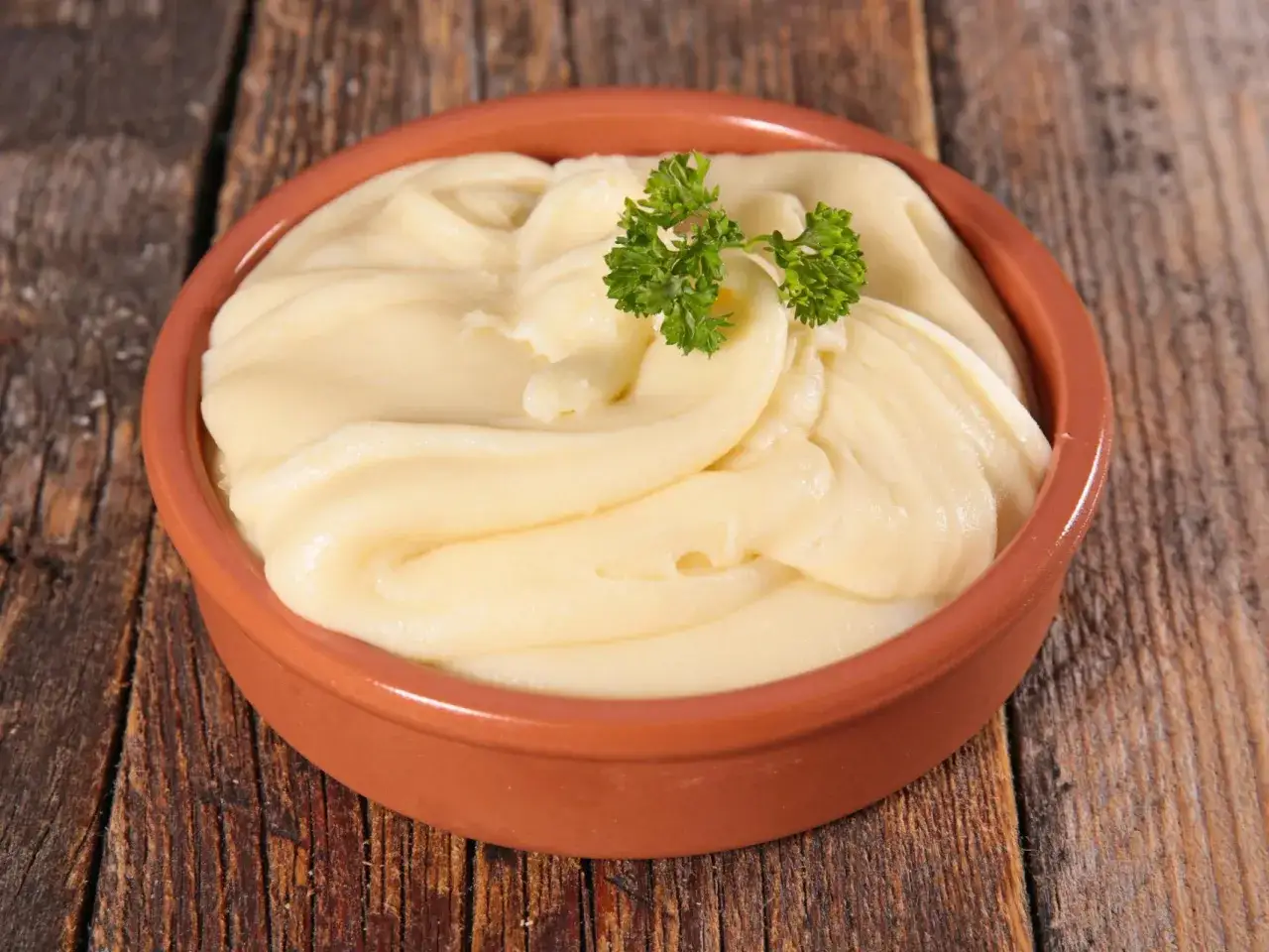 Un cuenco de barro con cremoso puré de patatas, adornado con una ramita de perejil fresco, sobre una mesa de madera rústica.