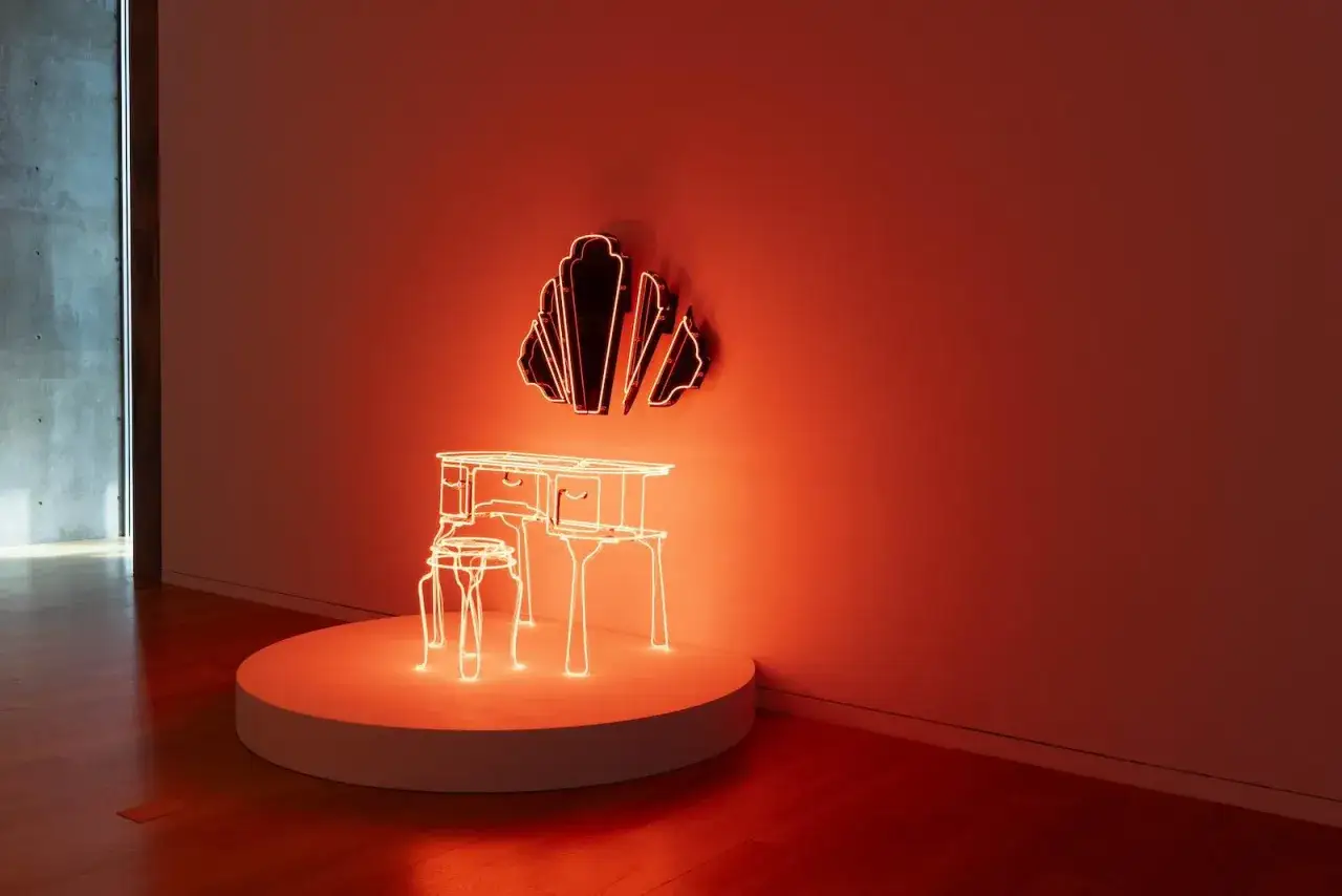Installation lumineuse évoquant le surréalisme : un bureau et une chaise en néon orange sur un socle, devant une forme murale déconstruite.