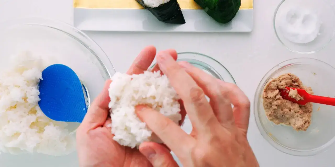 Hände formen Reis zu einem Ball, um Onigiri selber zu machen. Daneben Schüssel mit Thunfischfüllung und Salz.