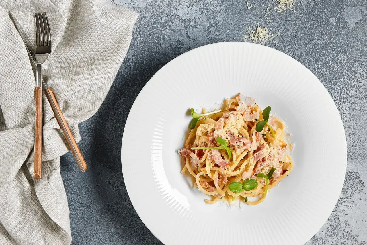 Assiette de tagliatelles carbonara cr&eacute;meuses, garnie de lardons et de pousses vertes, pr&ecirc;te &agrave; &ecirc;tre d&eacute;gust&eacute;e avec des couverts en cuivre.