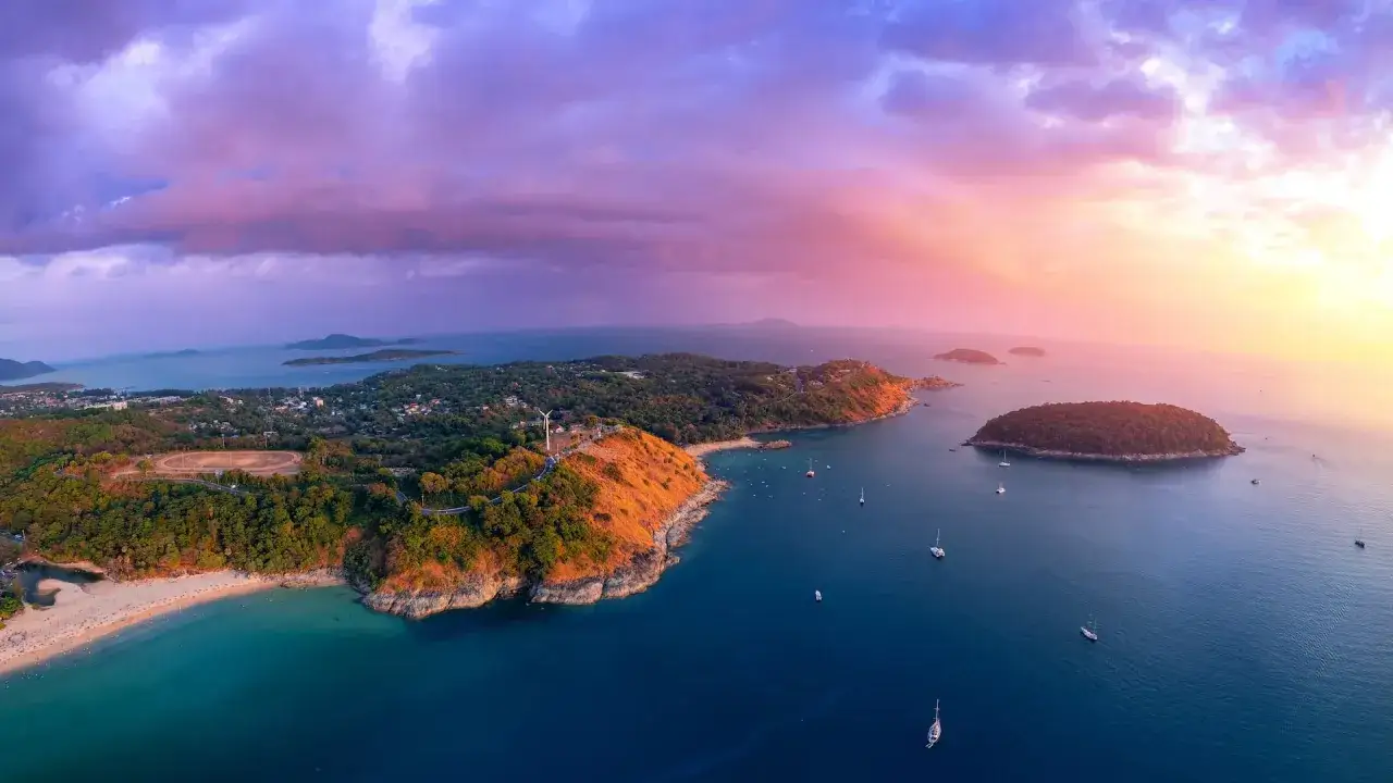 Zachód słońca nad wyspami i **plażami Phuket**. Turkusowa woda, zielone wzgórza i łodzie kołyszące się na falach.