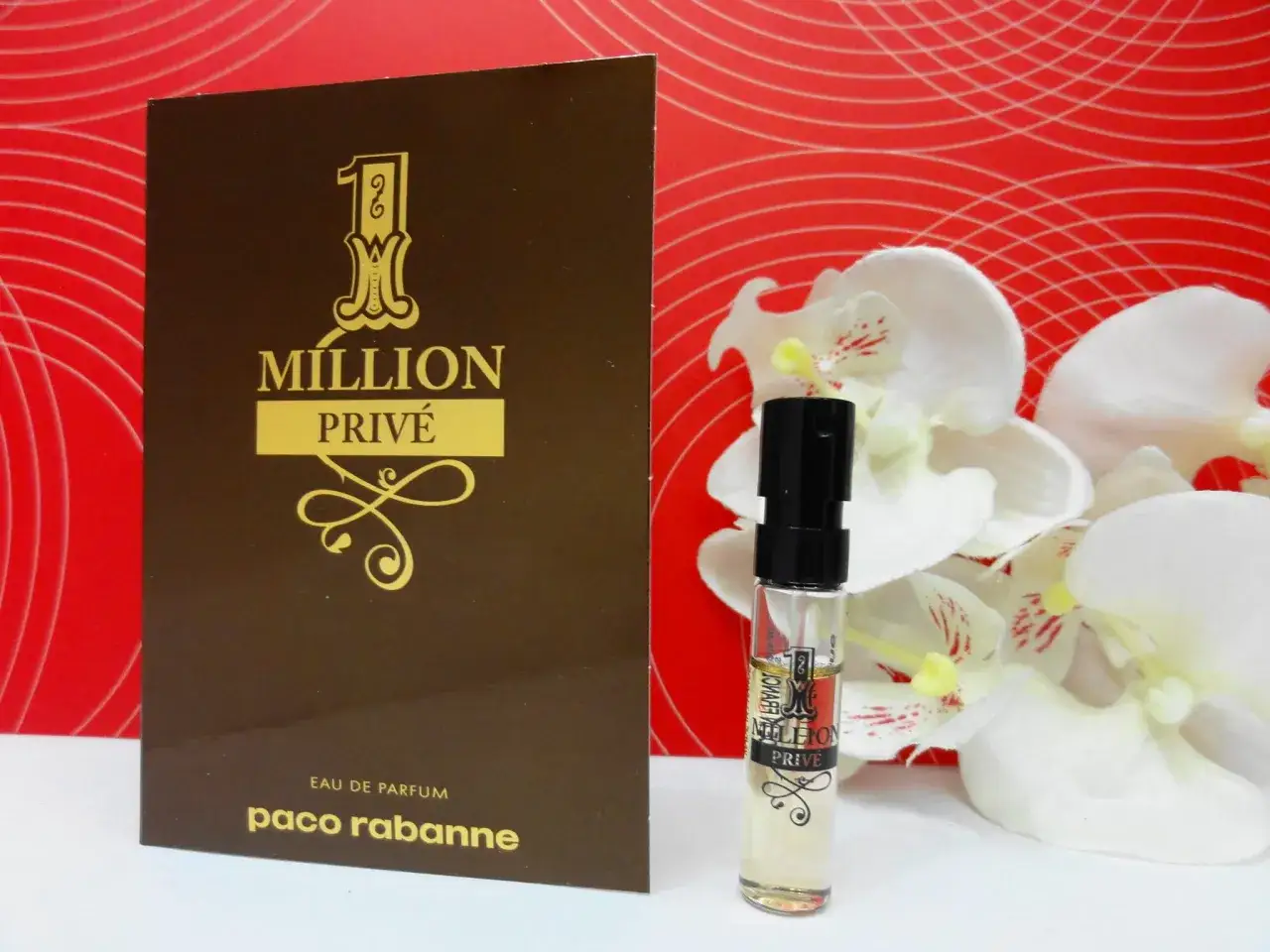 Złoty flakonik i opakowanie Paco Rabanne 1 Million Privé na tle czerwonych fal i białych kwiatów.