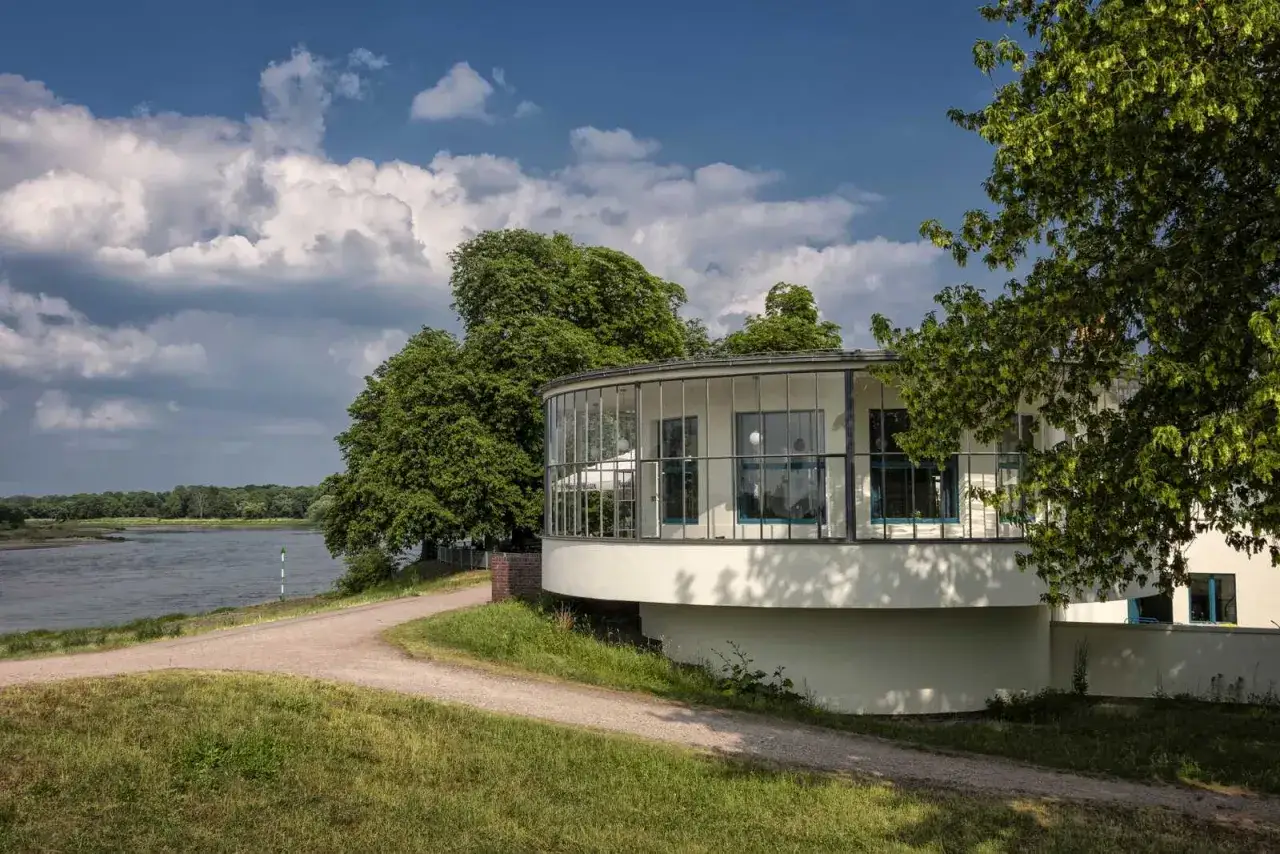 Runder Bau am Flussufer, ein Ort f&uuml;r Geheimtipps Dessau. Gr&uuml;ne B&auml;ume, blauer Himmel, Wolken.