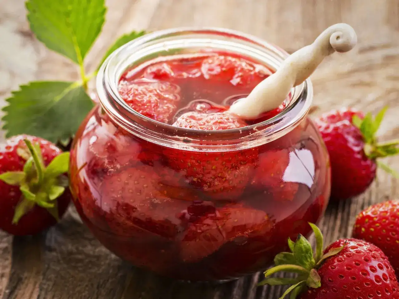 Ein Glas mit ganzen Erdbeeren in Sirup, bereit zum Einwecken. Frische Erdbeeren und ein grünes Blatt liegen daneben auf Holz.