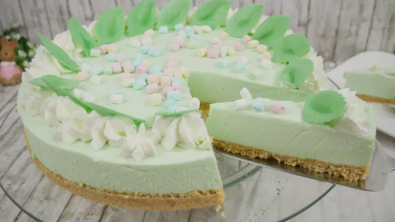 Eine helle, cremige Philadelphia Torte Waldmeister, verziert mit bunten Marshmallows und gr&uuml;nen Bl&auml;ttern. Ein St&uuml;ck wird gerade serviert.