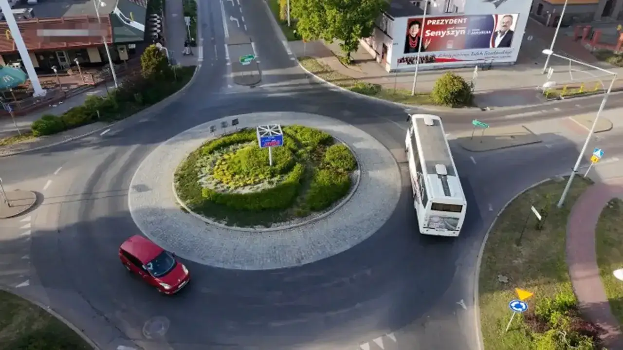Rondo z zieloną wyspą, czerwony samochód i biały autobus. W tle McDonald's i billboard. Brak sygnalizacji świetlnej, ruch kierowany znakami.
