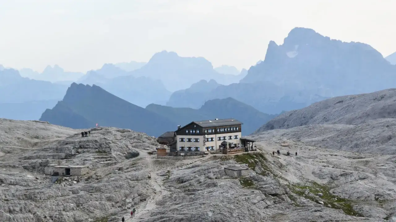 Il rifugio Rosetta, un edificio alpino con persiane blu, si erge su un terreno roccioso con montagne azzurre sullo sfondo.