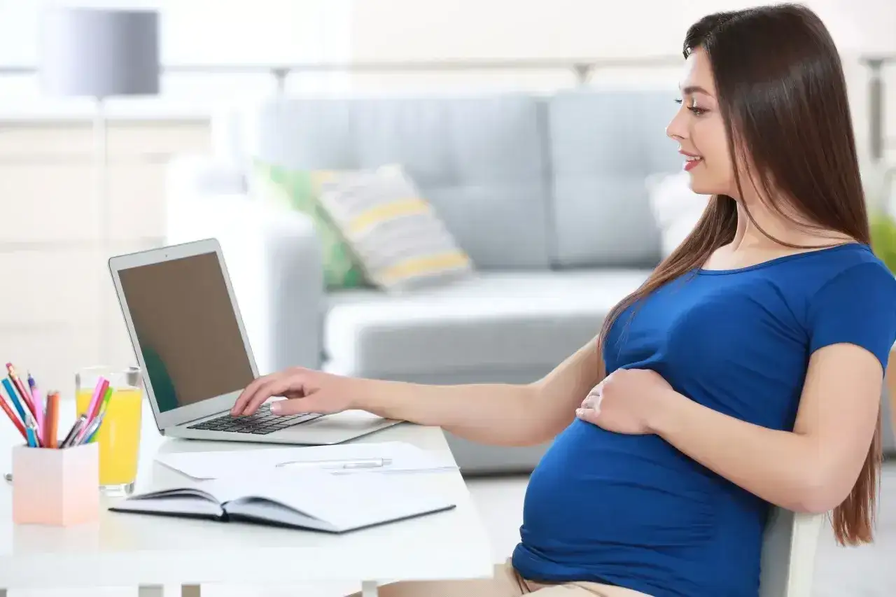 Uśmiechnięta, l4 w ciąży, pracuje przy laptopie w domowym biurze. Na biurku napój i przybory piśmiennicze.