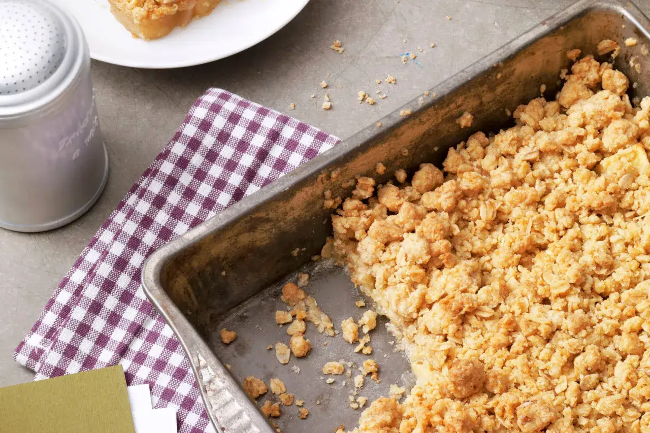 Ein frisch gebackener apfel crumble mit haferflocken in einer Metallform, bereit zum Servieren. Daneben liegt ein kariertes Tuch.