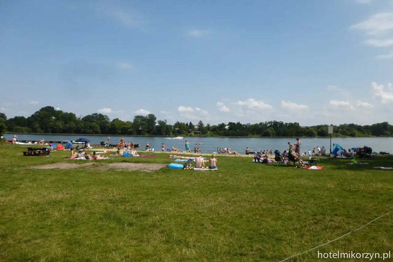 Ludzie odpoczywają na trawie i plaży nad jeziorem Mikorzyńskim. Woda jest spokojna, a na horyzoncie widać drzewa.