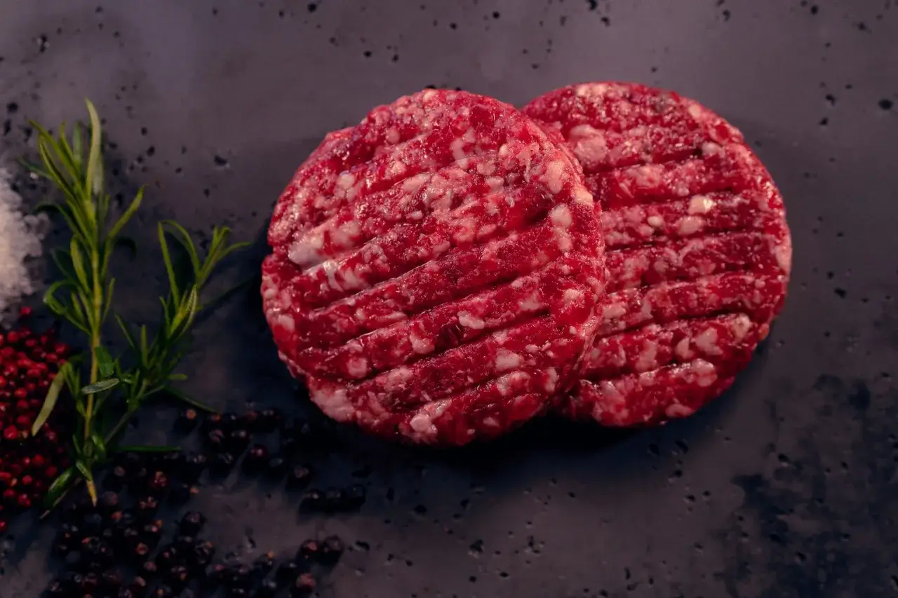 Dwa surowe kotlety wołowe, idealne na burgery, leżą obok gałązki rozmarynu i pieprzu. Zastanawiasz się, jakie mięso na burgery wybrać?