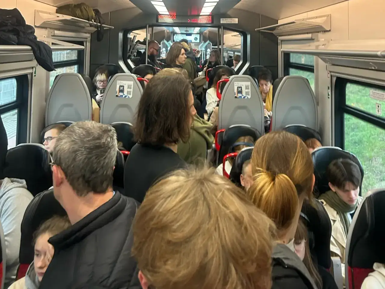 Wnętrze zatłoczonego pociągu relacji Poznań Nowy Tomyśl. Pasażerowie siedzą i stoją, niektórzy patrzą w telefony.