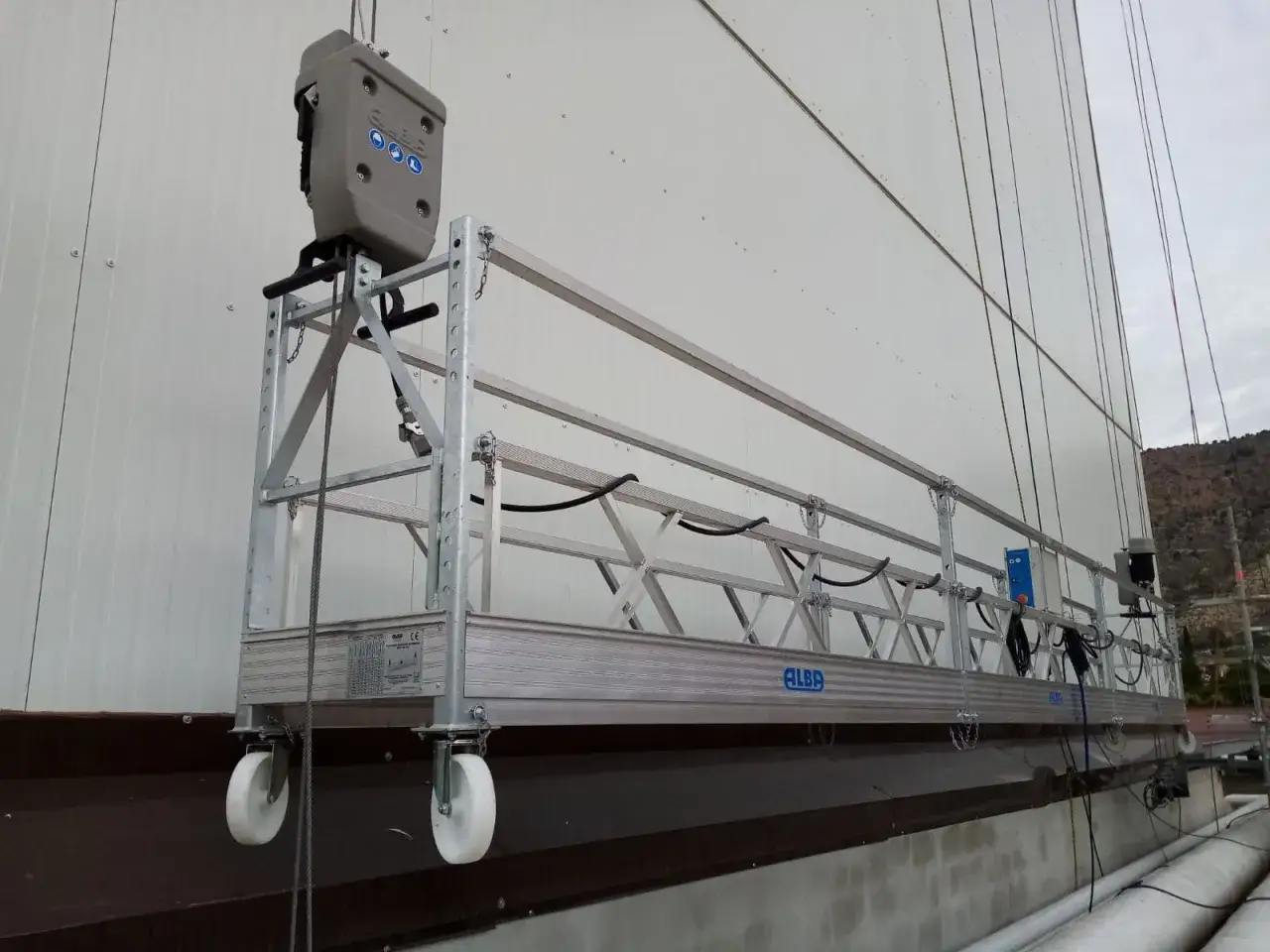 Andamios suspendidos listos para trabajar en altura. Plataforma metálica con ruedas y cables de seguridad.