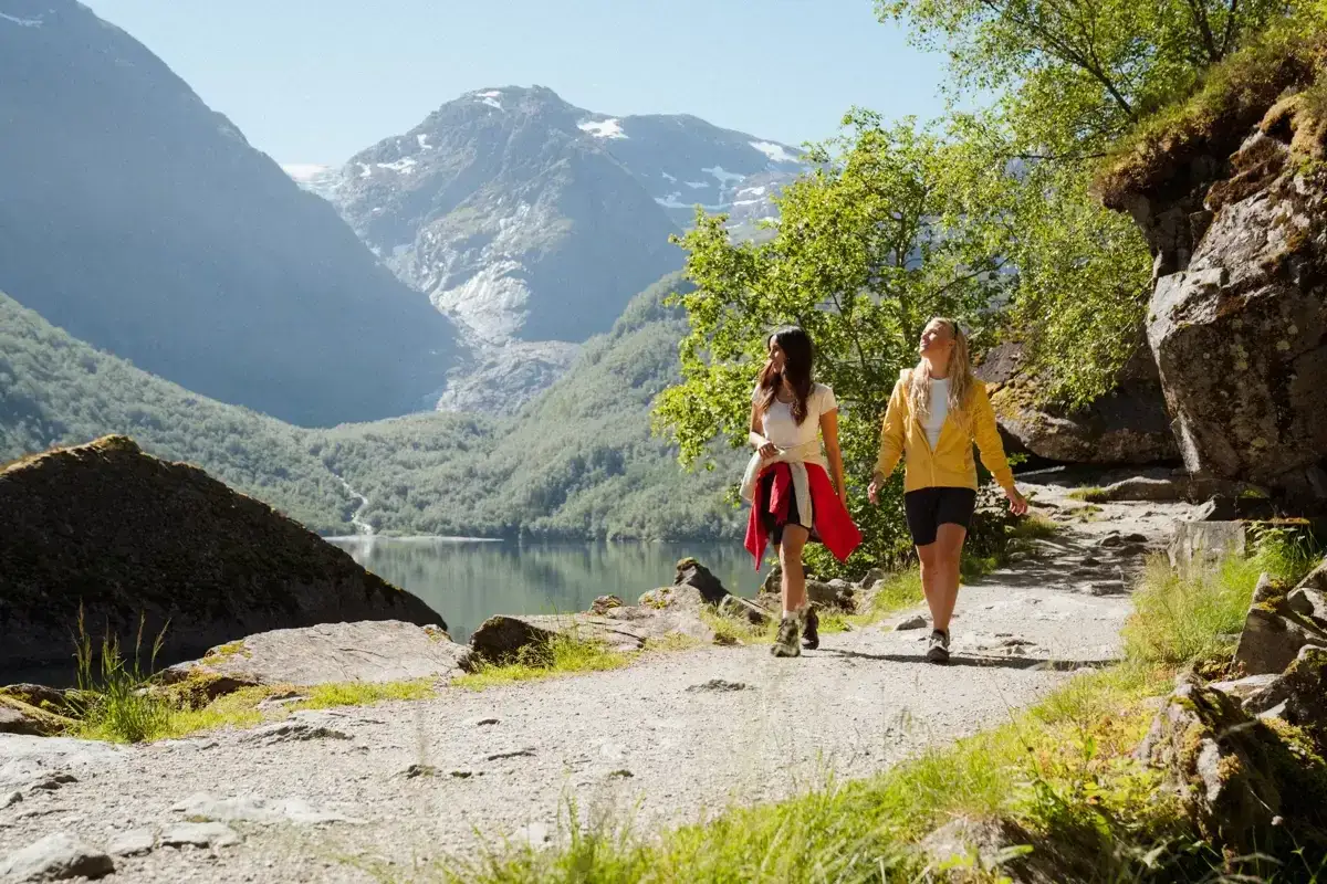 Tv&aring; kvinnor vandrar p&aring; en grusv&auml;g med en sj&ouml; och berg i bakgrunden. En perfekt dag f&ouml;r Hardangervidda vandring.