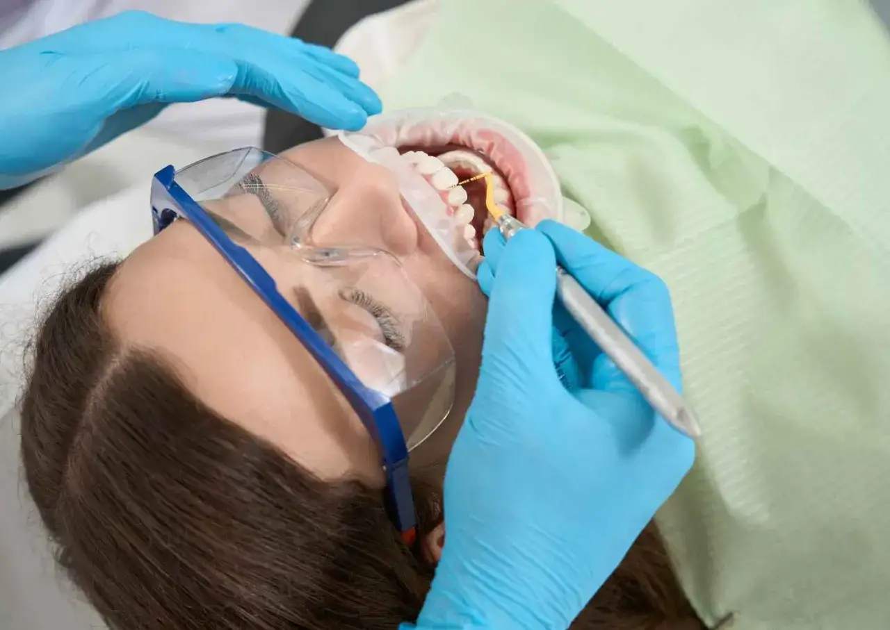 Dentysta wykonuje zabieg higienizacji zęb&oacute;w, usuwając kamień i osad. Pacjentka ma założone okulary ochronne i retraktory.