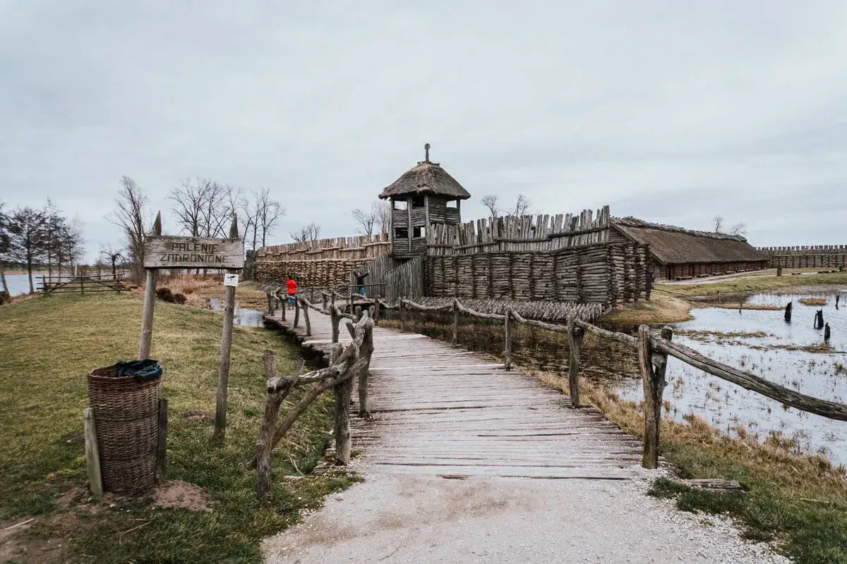 Drewniany mostek prowadzi do grodziska na szlaku piastowskim. Widać palisadę, wieżę strażniczą i chaty kryte strzechą.