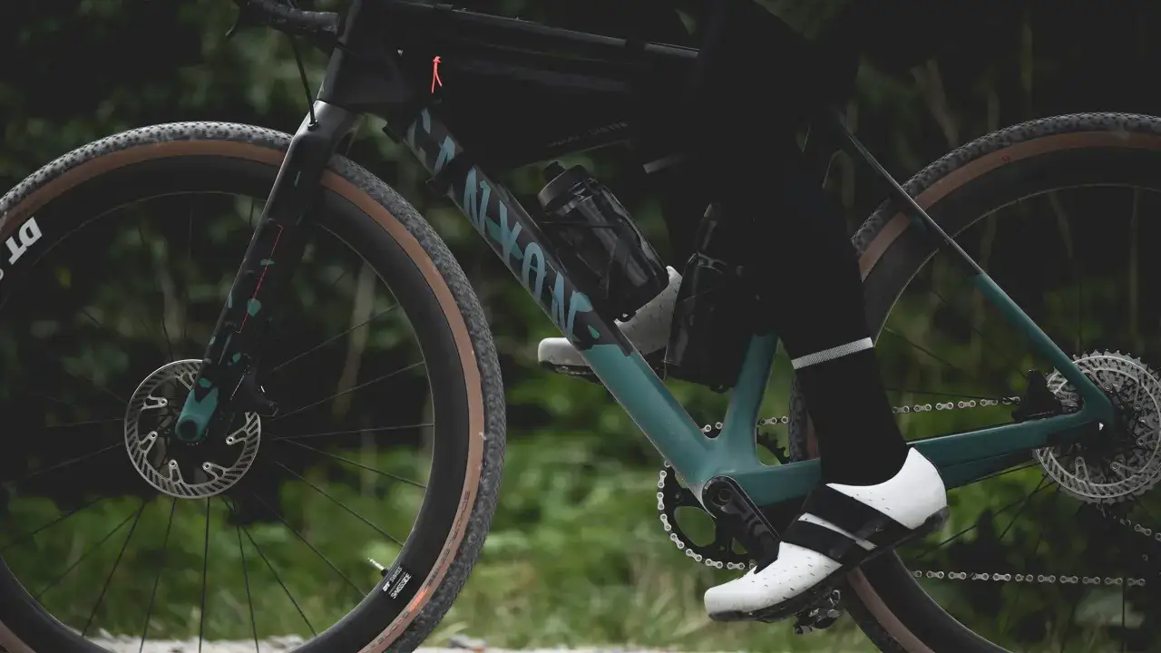 Rowerzysta w białych butach naciska na pedały do gravela. Widoczny rower z torbą podsiodłową i bidonami.