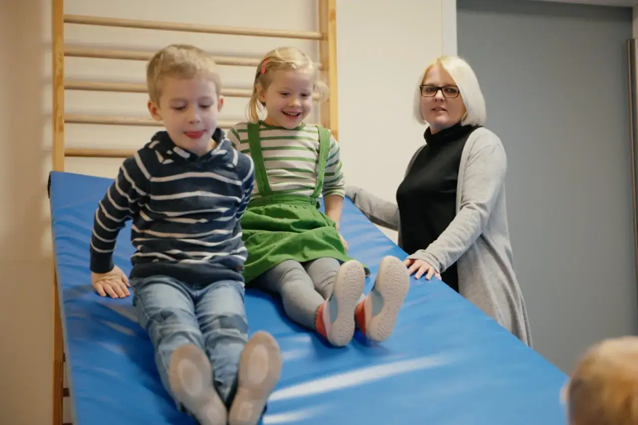 Zwei Kinder rutschen auf einer blauen Matte, während eine Erzieherin zusieht. Ein Fall könnte eine aufsichtspflichtverletzung kita darstellen.