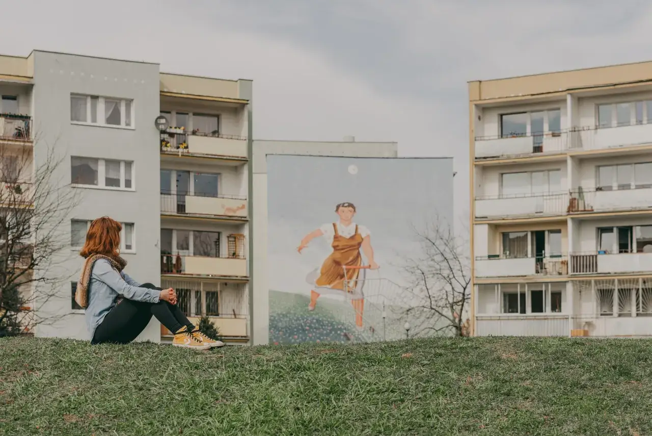 Dziewczyna na trawie podziwia mural z kobietą pchającą wózek. Nietypowe atrakcje Gdańsk.