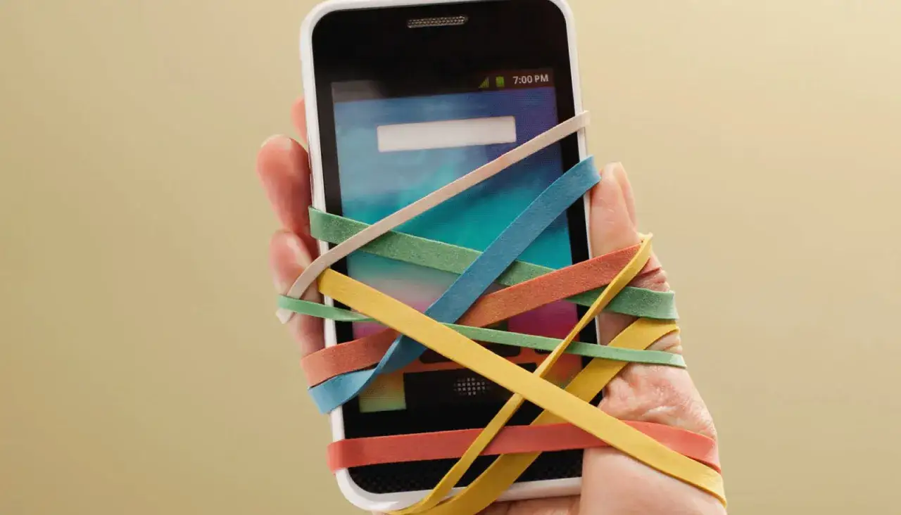 Mano sujetando un m&oacute;vil atado con gomas el&aacute;sticas de colores. Una met&aacute;fora visual sobre c&oacute;mo desengancharse del m&oacute;vil y recuperar el control.